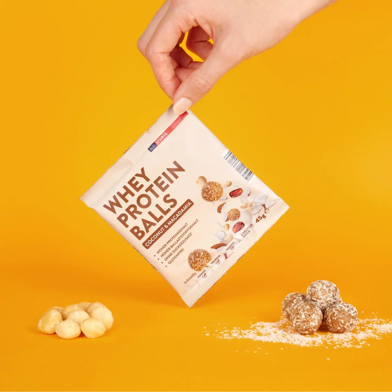 Energy Balls Coconut & Macadamia_04 Whey Protein Balls Coconut & Macadamia – Hand hält die Verpackung, Bälle und Zutaten daneben | Aldi Sports